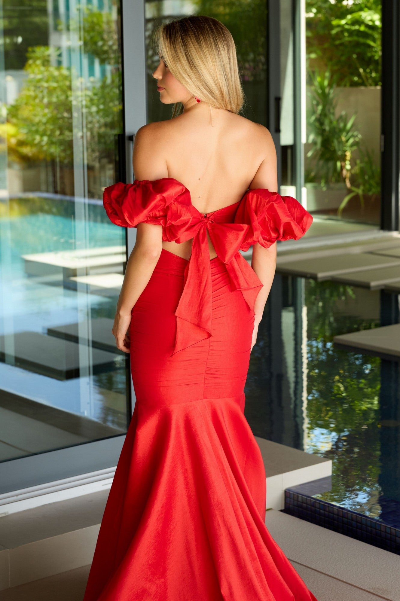 A woman in a red Long Prom Dress: Ava Presley 47172, featuring a strapless mermaid silhouette and large back bow, stands by a modern indoor pool facing away from the camera.
